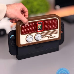 Puede incluir: Una base de Nintendo Switch marrón y crema con un diseño de radio retro. La base tiene una ranura para cartuchos de juego con un juego de Zelda insertado. Las palabras "Nintendo Switch" están impresas en la base.