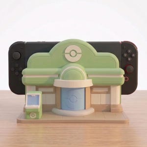 Puede incluir: Un soporte de consola de videojuegos verde y beige con forma de edificio, con una consola negra insertada. El soporte tiene un techo verde claro con un logotipo blanco, una puerta azul y una peque&ntilde;a m&aacute;quina verde. La consola tiene mandos rojos y negros.