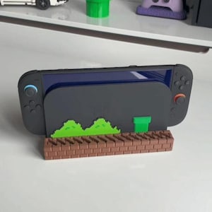 Puede incluir: Una consola de juegos portátil negra colocada en un soporte personalizado. El soporte presenta una base con patrón de ladrillos marrones, arbustos pixelados verdes y un diseño de tubería verde. La consola tiene mandos negros con un joystick azul y otro naranja.