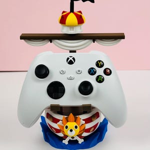 Peut inclure: Une manette Xbox blanche intégrée dans un design de navire décoratif. Le navire présente un drapeau noir, un mât jaune et rouge et une base bleue. Le design comprend un emblème de tête de mort et d'os croisés.