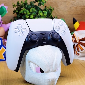 Puede incluir: Un mando de juego inalámbrico blanco y negro apoyado sobre un soporte con forma de cabeza esculpida, de color gris claro y textura rugosa. El mando incluye un panel táctil central, dos joysticks, una cruceta y cuatro botones de acción.
