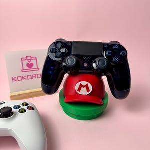 Può includere: Un controller per videogiochi blu scuro traslucido poggia su un supporto a forma di berretto rosso con una lettera M bianca, situato su una base circolare verde. Un controller bianco è parzialmente visibile in primo piano e un cartello dice KOKORO.