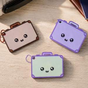 Puede incluir: Tres etiquetas de equipaje rectangulares con forma de maletas en miniatura, cada una con una cara de dibujos animados con grandes ojos negros y una boca pequeña. Una es beige con bordes marrones, una lavanda y una verde claro con borde morado.