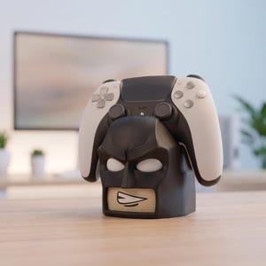 Pode incluir: Um comando de jogo preto e branco sobre um suporte preto com o tema Batman. O suporte tem a forma da cabeça da personagem com olhos brancos e uma zona da boca bege. O comando apresenta um design bicolor com pegas brancas.