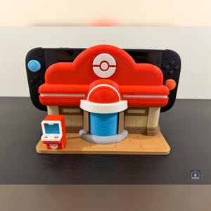 Puede incluir: Un accesorio rojo y blanco impreso en 3D, que se asemeja a un edificio con un logotipo circular, se coloca frente a una consola de juegos negra. A la izquierda hay una pequeña máquina de estilo arcade roja. El accesorio está diseñado para sostener la consola de juegos.