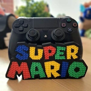 Può includere: Un supporto nero per controller di videogiochi con il logo Super Mario testurizzato e multicolore sulla parte anteriore. Il logo è composto da lettere in rilievo con motivo a maglia in blu, giallo, rosso e verde su una cornice nera.