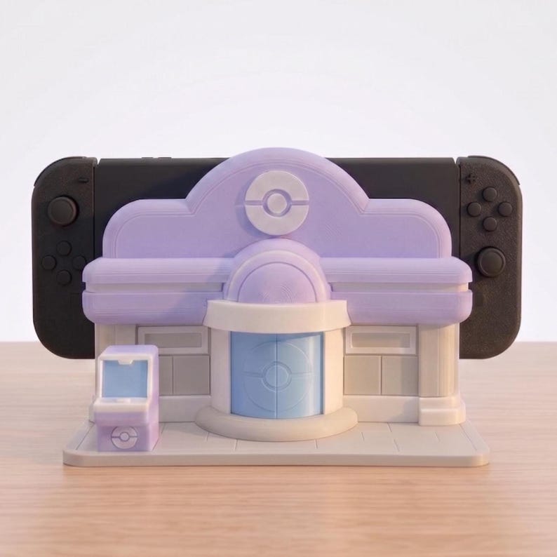 Puede incluir: Un soporte de consola de videojuegos morado y blanco con forma de edificio, con una peque&ntilde;a m&aacute;quina recreativa a juego. El soporte sostiene una consola de videojuegos negra. El edificio tiene un logotipo circular en el techo.