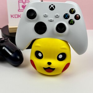 Op de afbeelding: Een witte draadloze videogamecontroller rustend op een gele, bolvormige karakterstandaard met zwarte ogen, een kleine neus en rode wangmarkeringen. De controller is voorzien van zwarte analoge sticks, een D-pad en vier gekleurde actieknoppen.