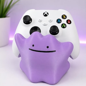 Könnte beinhalten: Ein violetter, klecksförmiger Controller-Ständer mit einem einfachen Gesicht, bestehend aus zwei schwarzen Punkten als Augen und einem dünnen schwarzen Strich als Mund. Der Ständer hält einen weißen kabellosen Gamecontroller mit schwarzen Joysticks.