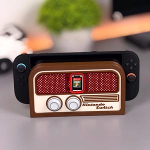 Puede incluir: Un soporte para cartuchos de juego Nintendo Switch en color marrón y crema diseñado como una radio antigua. El soporte cuenta con una rejilla de altavoz roja, dos perillas circulares blancas y el texto Nintendo Switch. Incluye un cartucho de Zelda.
