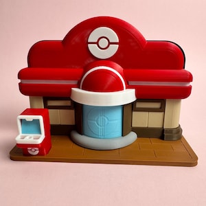 Puede incluir: Modelo impreso en 3D de un Centro Pokémon, con techo rojo y emblema de Poké Ball blanco. El edificio tiene una puerta azul, una máquina expendedora roja y una base marrón. El modelo es detallado.