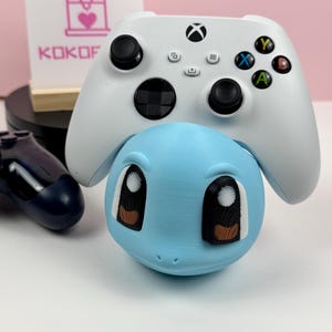 Op de afbeelding: Een lichtblauwe 3D-geprinte controllerstandaard in de vorm van een karakterhoofd met grote ogen en bruine pupillen, die een witte draadloze gamecontroller met zwarte joysticks en gekleurde knoppen ondersteunt. Een bordje met KOKOF is zichtbaar.