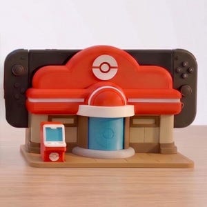 Puede incluir: Un dock para Nintendo Switch con forma de edificio rojo y blanco con una puerta azul y una peque&ntilde;a m&aacute;quina recreativa roja. El dock est&aacute; dise&ntilde;ado para parecerse a un Centro Pok&eacute;mon, con una consola Nintendo Switch negra.