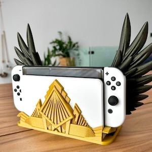 Può includere: Una console di gioco bianca con comandi neri e un supporto color oro. Il supporto presenta un design Art Déco e ali nere si estendono dai lati della console. La console è progettata per contenere un dispositivo di gioco portatile.