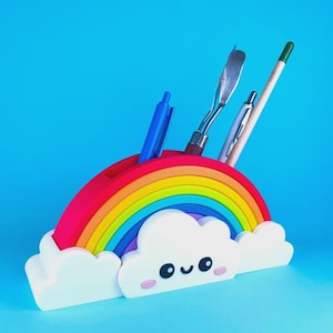 Può includere: Un organizer da scrivania a forma di arcobaleno con una base a nuvola bianca che presenta un volto sorridente, guance rosa e occhi neri. L'organizer ha diverse fessure che contengono una penna blu, un utensile in metallo, una penna argentata e una matita verde.