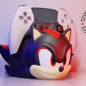 Op de afbeelding: Een karakter-thema PlayStation 5 controllerhouder in zwart, rood en crème. De houder is ontworpen om op een personage met een lachend gezicht te lijken. Een witte en zwarte controller is in de houder geplaatst.