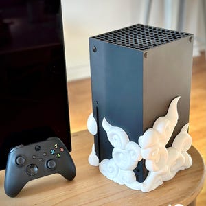 Puede incluir: Consola Xbox Series X negra con una base decorativa blanca en forma de nube. Un mando Xbox negro está a la izquierda. La consola está sobre una mesa de madera. El texto "HOLO PROPS" es visible.