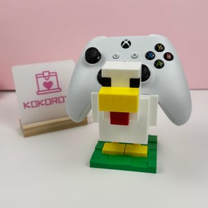 Puede incluir: Un soporte para mando de videojuegos blanco con forma de pollo cúbico, con pico amarillo, barbilla roja y ojos negros, sobre una base verde. Al lado hay un cartel blanco con un icono de impresora 3D rosa y el texto KOKORO impreso en color rosa.