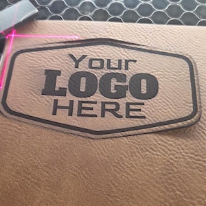 May include: A laser engraving machine in operation, focused on a tan leather surface. The machine is etching a logo that reads "Your LOGO HERE" within a dark-lined, shield-shaped border. The laser's red beam is visible, highlighting the engraving process.