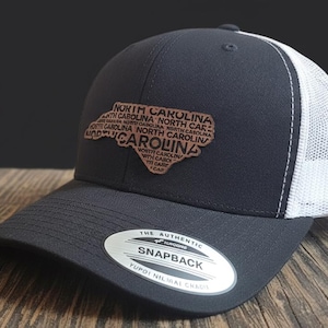 Op de afbeelding: Een marineblauwe en witte truckerpet met een bruine leren patch in de vorm van North Carolina en de naam van de staat. De pet heeft een gebogen klep en een snapback-sluiting.