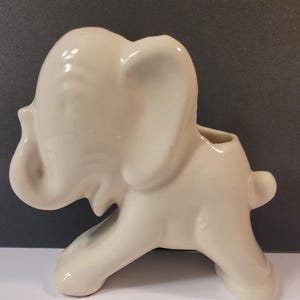 May include: Ivory-colored ceramic elephant figurine. The elephant has a trunk, ears, and a small tail. The figurine is in a walking pose and has a small opening on its back. This decorative piece is suitable for home decor.