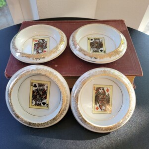 May include: Four white porcelain dishes with gold rims and playing card images in the center. The playing card suits are hearts, clubs, diamonds, and spades. The dishes are arranged on a dark surface.