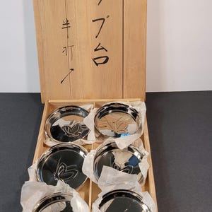 May include: A set of six silver-colored coasters with a butterfly design, nestled in a wooden box. The box has Japanese characters on the front. Each coaster is approximately 4 inches in diameter.