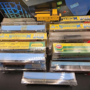 May include: A collection of N scale model train cars in clear plastic cases, including a yellow caboose, a Tech II Railpower 1400 power pack, and various freight cars with Minitrix and Model Power branding, all arranged in a stacked display.