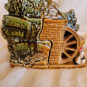 May include: A ceramic decorative piece depicting a water mill scene. The piece is primarily brown and green, with a brick-like texture on the mill and a water wheel. The text "Down by the old Mill Stream" is visible on a banner.