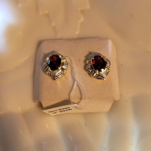 May include: A pair of gold-toned stud earrings with a central, deep red gemstone surrounded by smaller, clear stones. The earrings are displayed on a small, off-white card. The tag reads "MADE IN THAILAND."