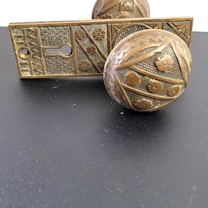May include: An antique bronze door handle set, comprising two oval doorknobs and a rectangular backplate. The doorknobs and backplate are decorated with floral and geometric patterns. The set is designed for a door and is made of metal.