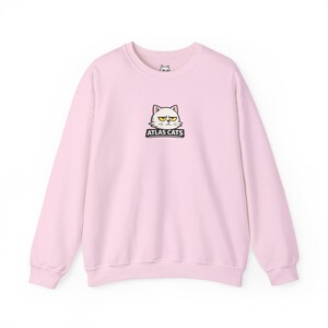 May include: A light pink sweatshirt with a crew neck. The front features a graphic of a grumpy cat face with the text "ATLAS CATS" in a black rectangle. The sweatshirt is made of a soft material.