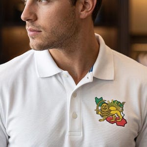 Puede incluir: Polo blanco con tapeta de tres botones y un pequeño emblema bordado con un diseño de león y sol. La camisa tiene cuello clásico y mangas cortas. El emblema es verde, rojo, amarillo y blanco.