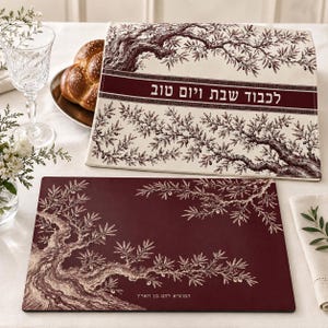 May include: A rectangular challah cover in cream fabric with a dark red olive tree illustration and Hebrew text, paired with a matching dark red rectangular mat featuring a cream olive tree design and Hebrew text at the bottom edge.