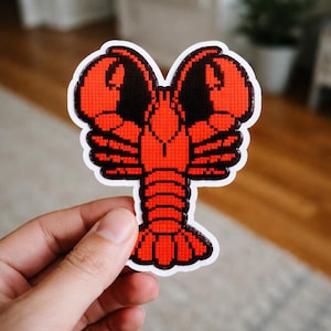 May include: A red and black pixelated lobster sticker with a white border. The sticker features a detailed design of a lobster with its claws raised. The sticker is held by a hand.