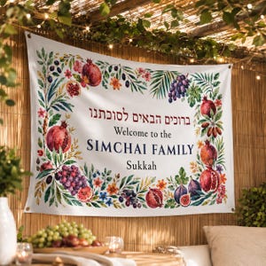 May include: A white rectangular banner featuring a watercolor border of pomegranates, grapes, figs, and palm leaves. The text reads: "ברוכים הבאים לסוכתנו", "Welcome to the SIMCHAI FAMILY", and "Sukkah". The banner has metal grommets in the corners.