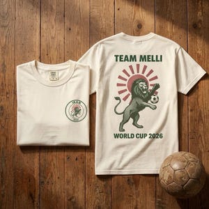 May include: Off-white short-sleeve t-shirt featuring a green and red graphic of a lion playing soccer. The back displays the text TEAM MELLI and WORLD CUP 2026. The front chest area has a circular logo with the text IRAN and 2026.