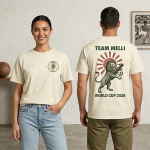 May include: An off-white short-sleeve t-shirt featuring a small circular logo on the front chest. The back displays a large graphic of a lion playing soccer in front of a red sunburst, with the text TEAM MELLI and WORLD CUP 2026 printed in green.