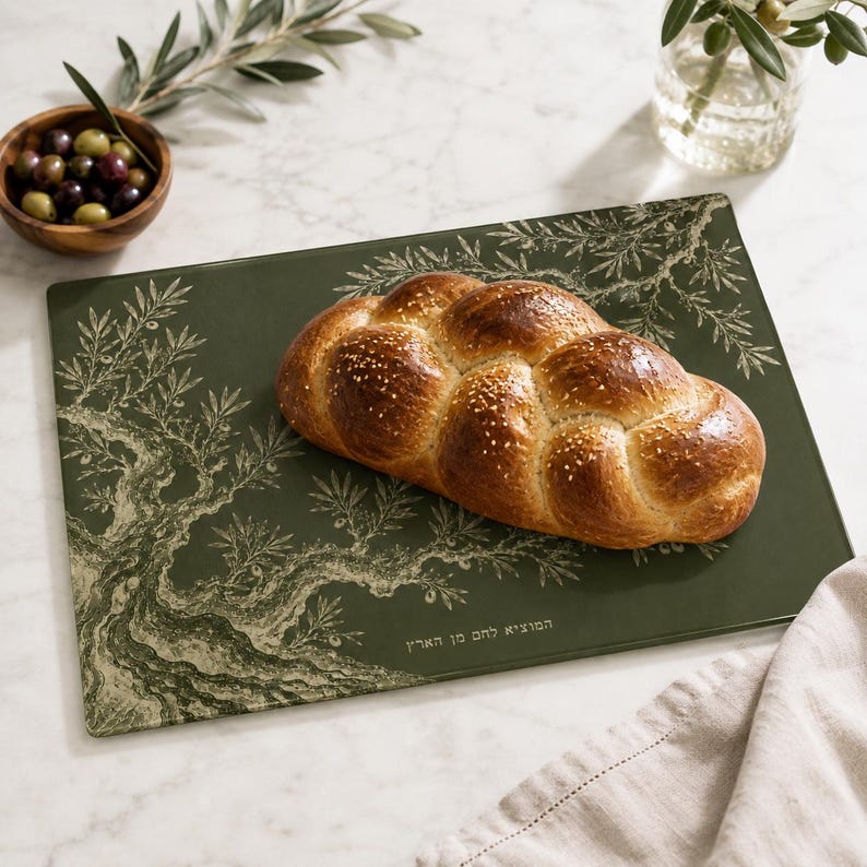 May include: A rectangular dark green cutting board featuring a detailed illustration of an olive tree and Hebrew text. A golden-brown braided challah bread topped with sesame seeds rests on the surface of the board.
