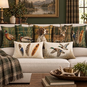 May include: A collection of seven rectangular throw pillows featuring wildlife and nature themes. The designs include a pheasant, a mallard duck, a hunting dog, a deer, and various bird feathers, set against green plaid and cream-colored backgrounds.