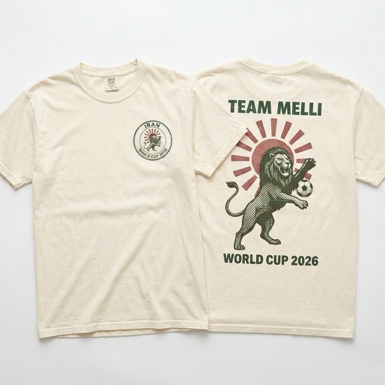 May include: A cream-colored short-sleeve t-shirt featuring a small circular logo on the front chest with a lion and the text IRAN WORLD CUP 2026. The back displays a large graphic of a lion playing soccer with the text TEAM MELLI and WORLD CUP 2026.
