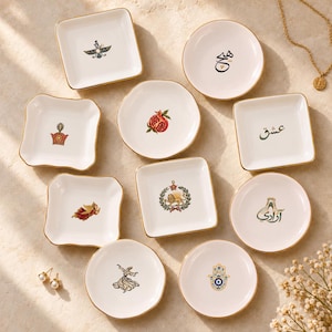 May include: A collection of ten white ceramic trinket dishes with gold-trimmed edges, featuring various illustrations including a pomegranate, a lion, a hamsa, a dervish, and Persian calligraphy, arranged on a light-colored surface.