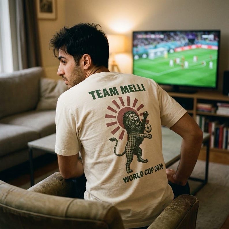May include: A cream-colored t-shirt featuring a graphic on the back with the text "TEAM MELLI" and "WORLD CUP 2026" in green. The design includes a stylized dark green lion holding a soccer ball in front of a red sunburst pattern.