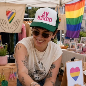 May include: A white and green trucker hat with the text "QUEER AF" in pink. The hat is worn by a person with sunglasses and tattoos. A rainbow flag and other pride-themed items are visible in the background.