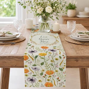 May include: A floral table runner with a cream background, featuring orange, yellow, and purple flowers. The runner has the text "Love Grows Here" in a circular design. The table is set with plates, glasses, and a vase of flowers.