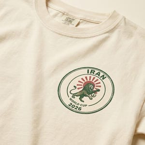 May include: An off-white t-shirt featuring a circular graphic on the chest. The design includes a green lion, a red sunburst, and a soccer ball, with the text IRAN WORLD CUP 2026 printed in green inside a dark green circular border.