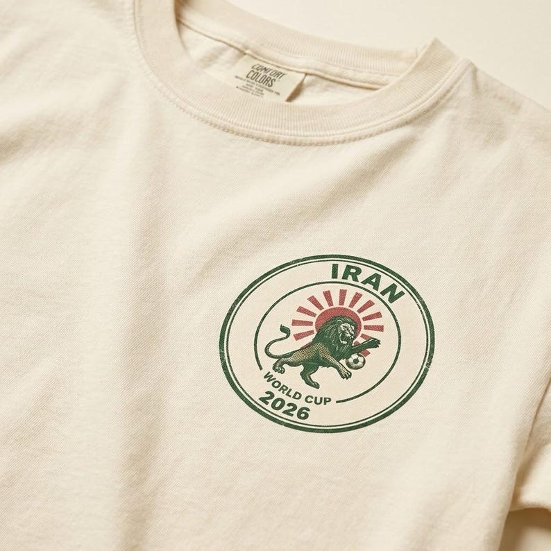May include: An off-white t-shirt featuring a circular graphic on the chest. The design includes a green lion, a red sunburst, and a soccer ball, with the text IRAN WORLD CUP 2026 printed in green inside a dark green circular border.