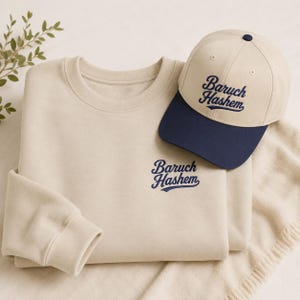 Puede incluir: Una sudadera beige de cuello redondo y una gorra de béisbol a juego con visera azul marino. Ambos artículos presentan el texto Baruch Hashem bordado en letra cursiva azul marino. El texto aparece en el pecho de la sudadera y en la gorra.