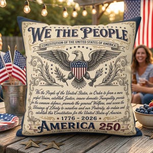 Pode incluir: Uma almofada decorativa com um design patriótico. A almofada apresenta o texto "We The People" e uma ilustração de uma águia. A almofada também inclui o texto "America 250" e os anos 1776-2026.