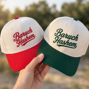 Puede incluir: Dos gorras de béisbol con coronas color blanco hueso y el texto bordado Baruch Hashem. Una gorra tiene visera y letras rojas, mientras que la otra tiene visera y letras verde oscuro. Ambas gorras tienen un botón en la parte superior.